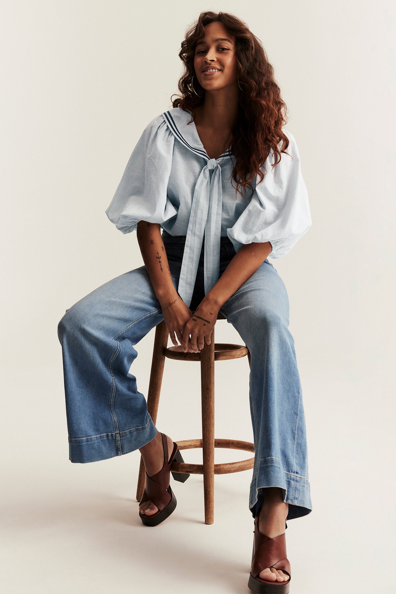Front view of a woman sitting in a light blue striped sailor blouse, wide-leg jeans, and brown sandals.