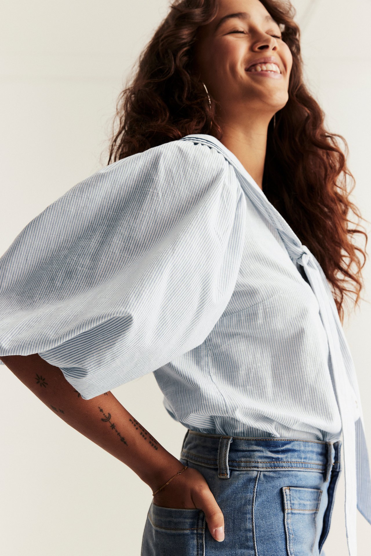 Woman in light blue striped cotton sailor blouse with puff sleeves and tie, and blue jeans.