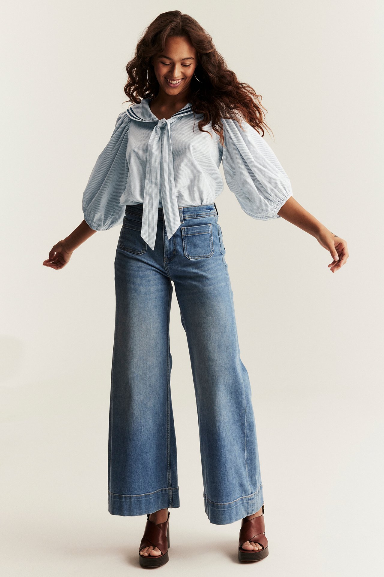 Woman wears light blue striped sailor blouse with tie, puff sleeves, blue wide-leg jeans, brown platform sandals.
