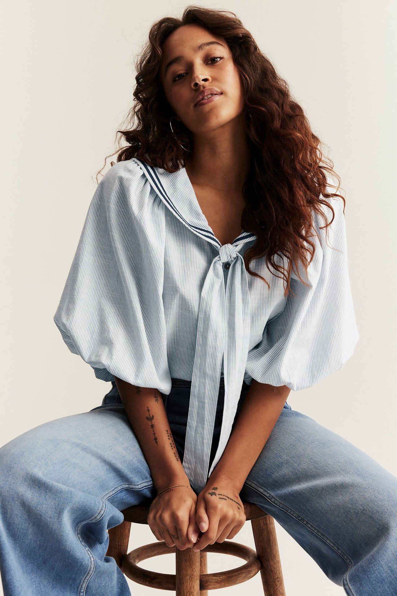 Front view of a woman wearing a light blue striped woven sailor blouse with puff sleeves and tie details, paired with wide-leg jeans.