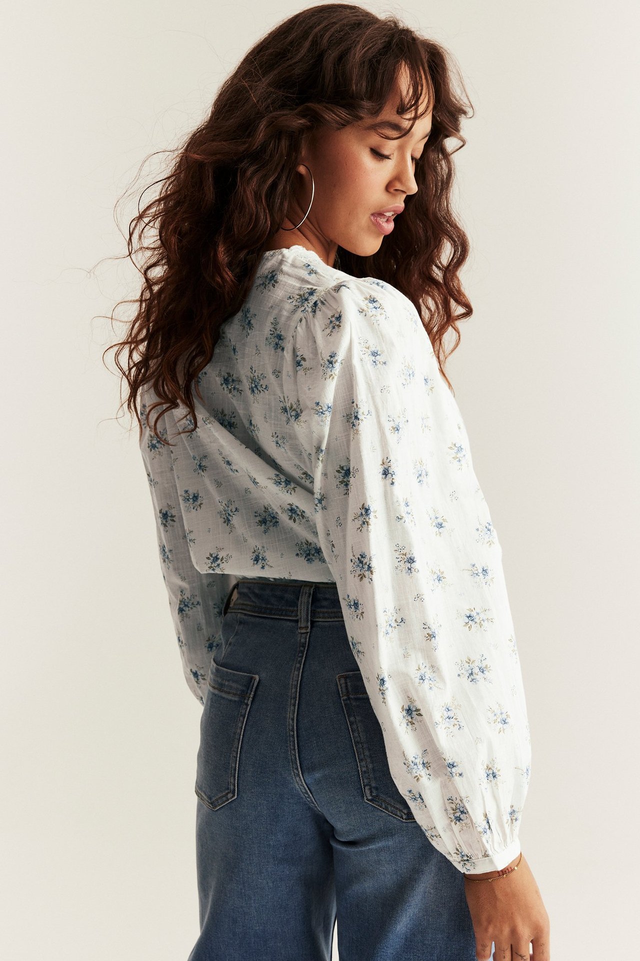 Side view: Woman in offwhite floral cotton puff-sleeve blouse, blue jeans, gold hoop earrings.