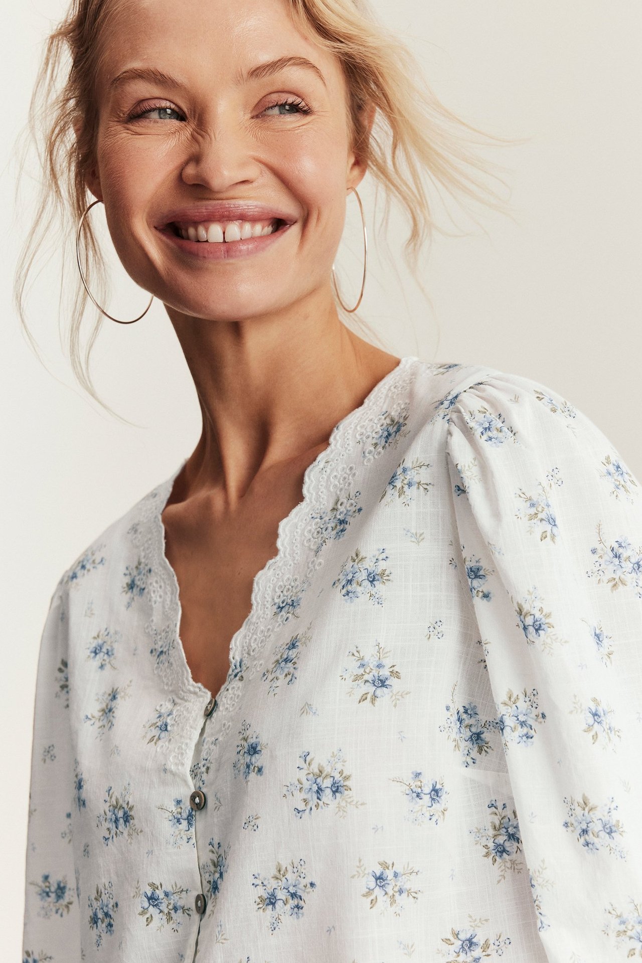 Nahaufnahme: Frau in offwhite Blumen-Baumwollbluse mit besticktem V-Ausschnitt, Puffärmeln und goldenen Kreolen.