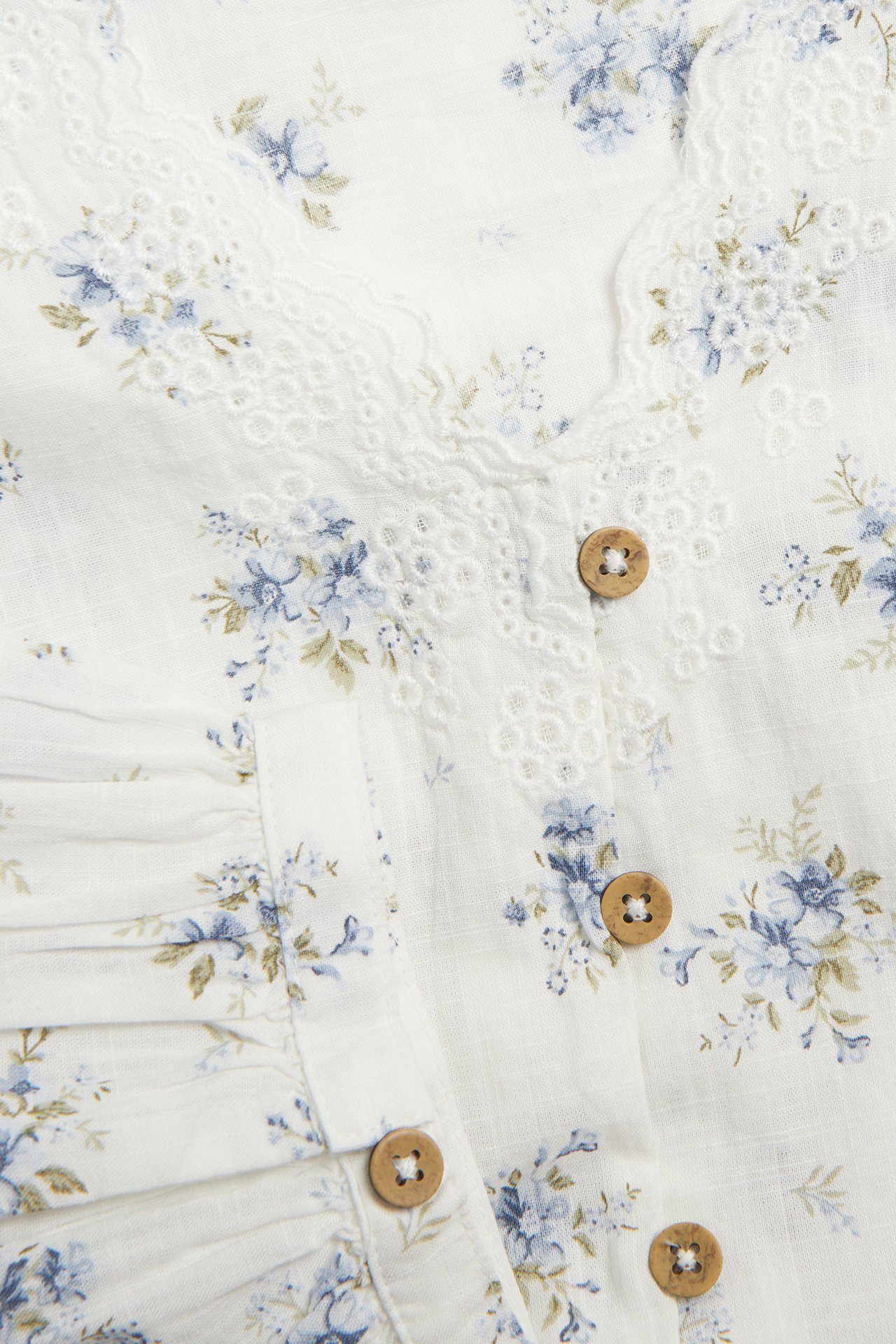 Nahaufnahme einer cremeweißen floralen Baumwollbluse mit besticktem V-Ausschnitt und Holzknöpfen.