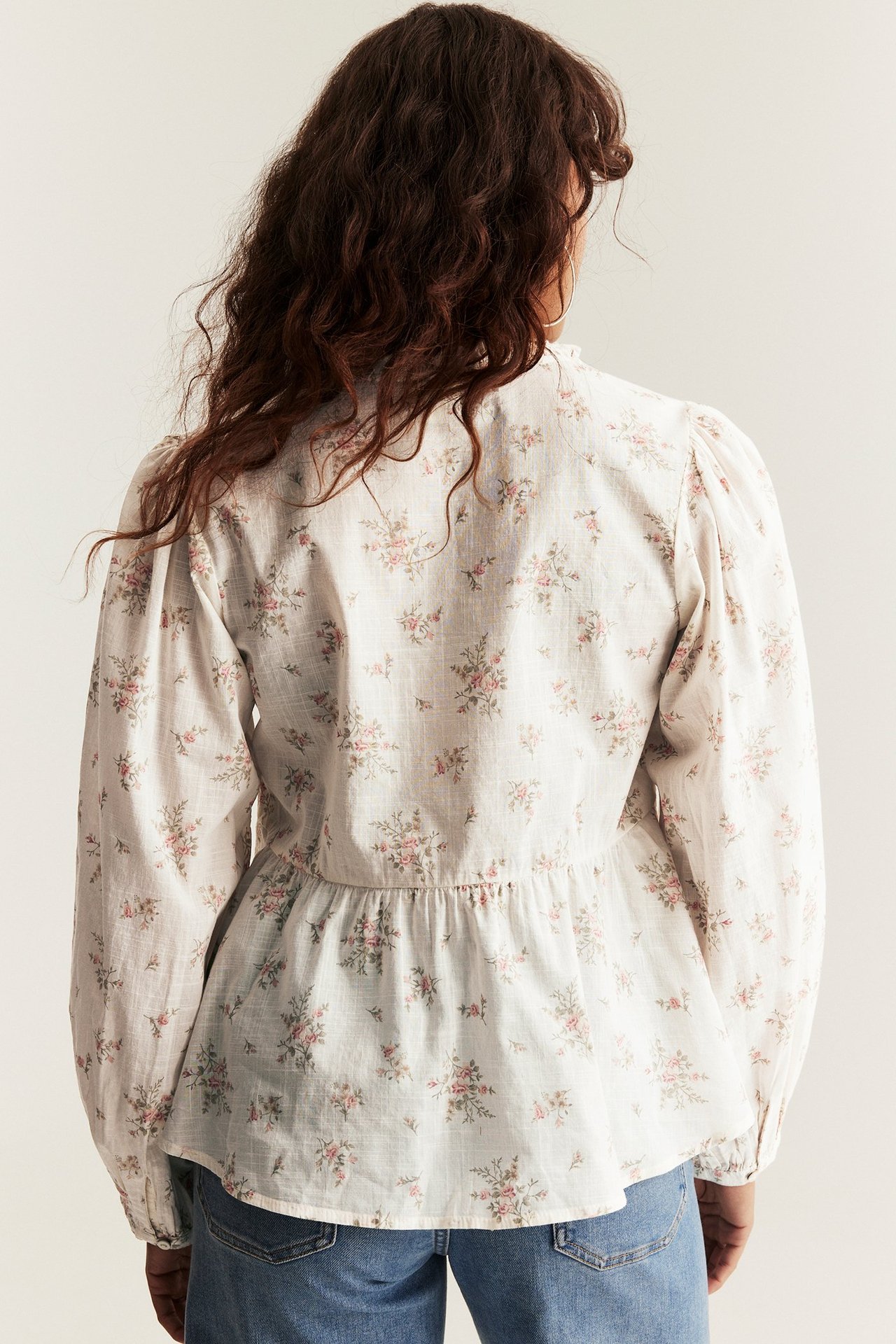 Back view: Woman wears an off-white floral cotton peplum blouse with long sleeves and blue jeans.