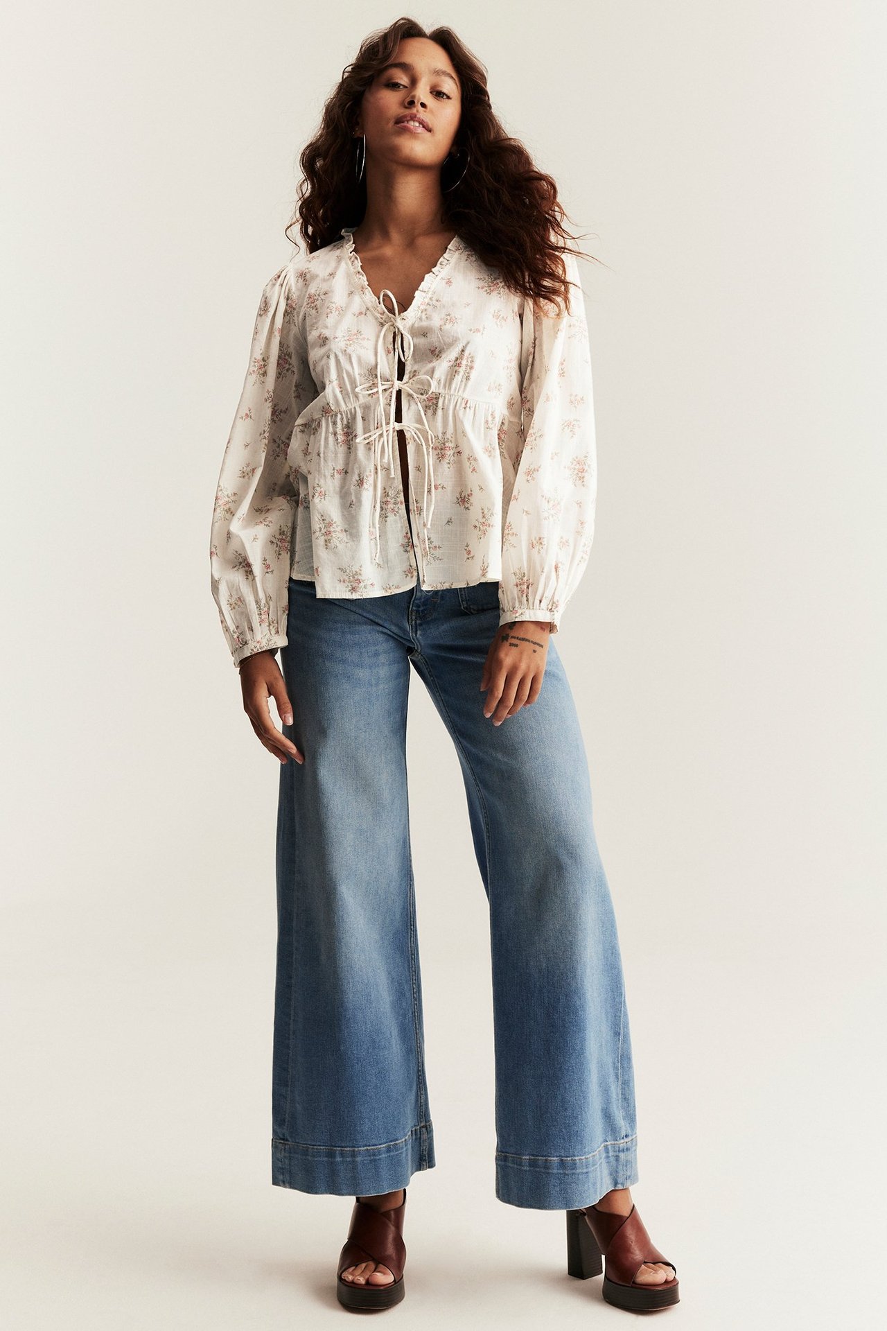 Front view of a woman in an offwhite floral cotton blouse with tie-front, light blue jeans, brown platform sandals.