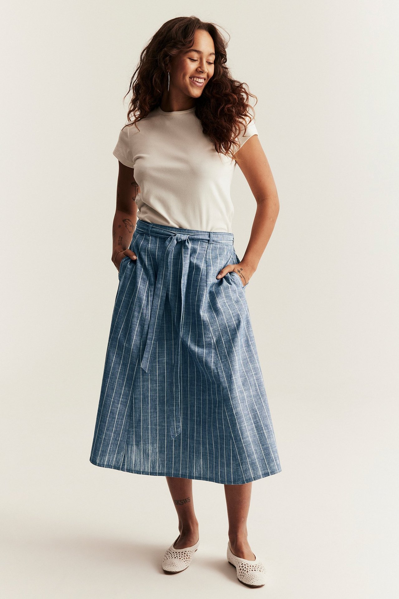 Front view: Woman in blue striped midi skirt with belt, white t-shirt, and cream perforated flats.