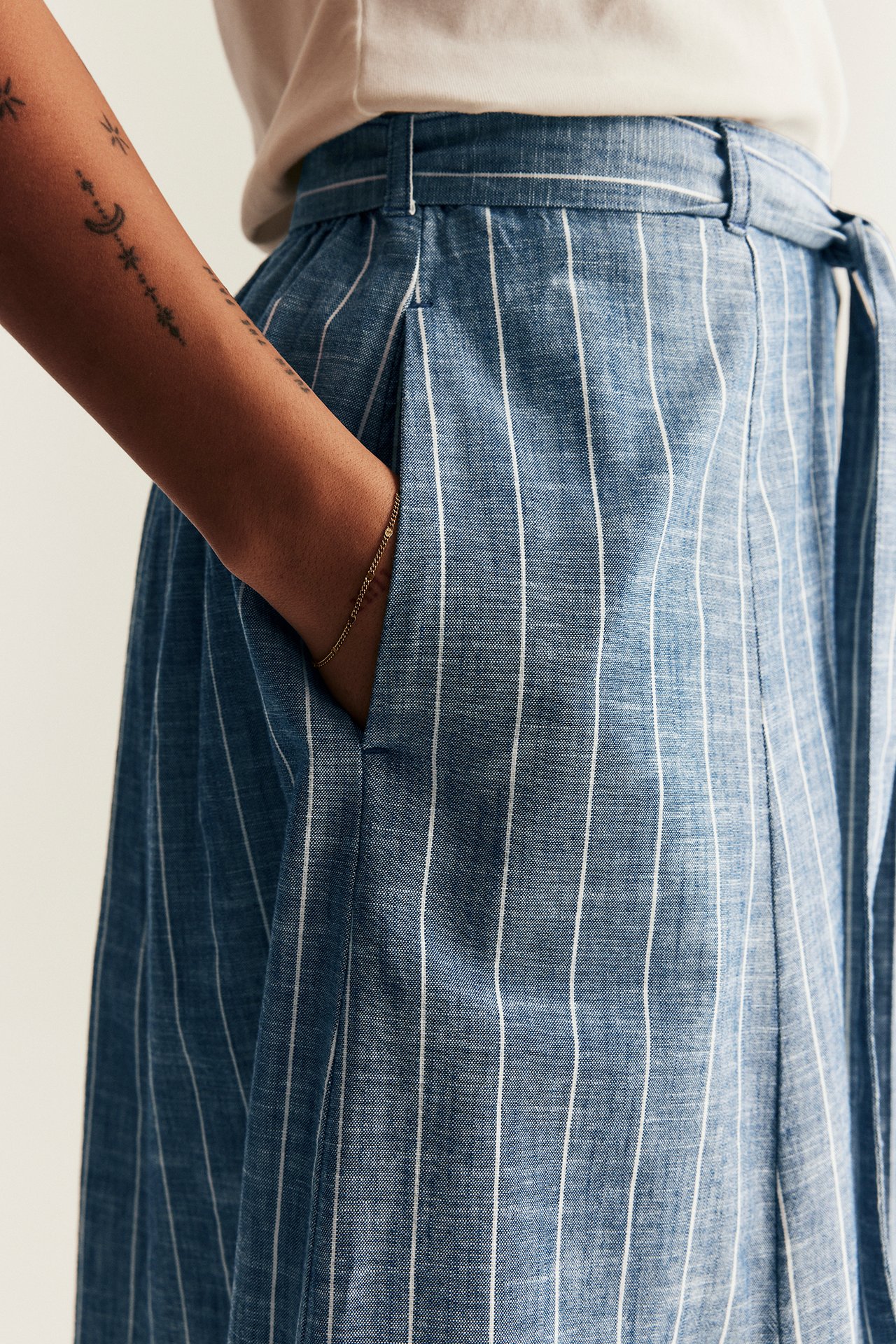 Close-up of a woman in a blue striped woven skirt with side pocket, tie belt, and a light t-shirt and gold bracelet.