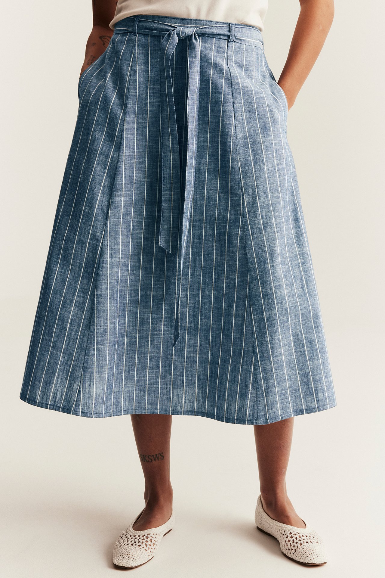Front view: Woman in blue striped cotton midi skirt with tied belt, pockets, and white crocheted flats.