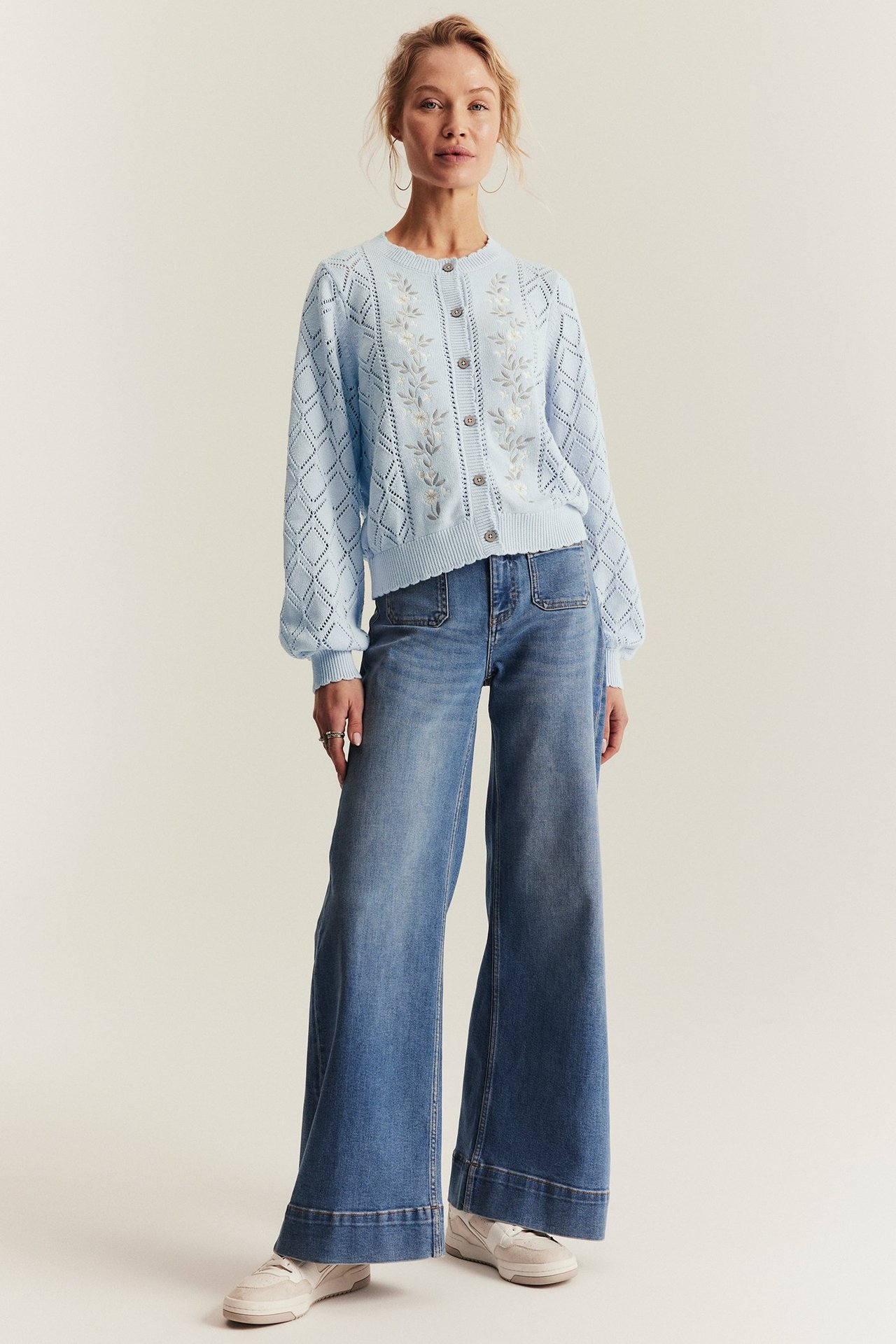 Front view of a woman wearing a light blue embroidered knitted cotton cardigan, wide-leg blue jeans, and white sneakers.