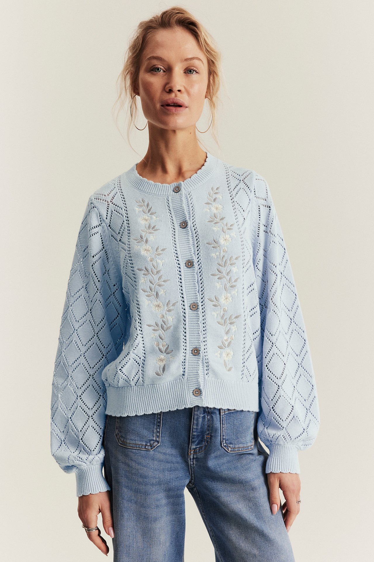 Front view: Woman in a light blue embroidered cotton cardigan with diamond knit sleeves and blue jeans.