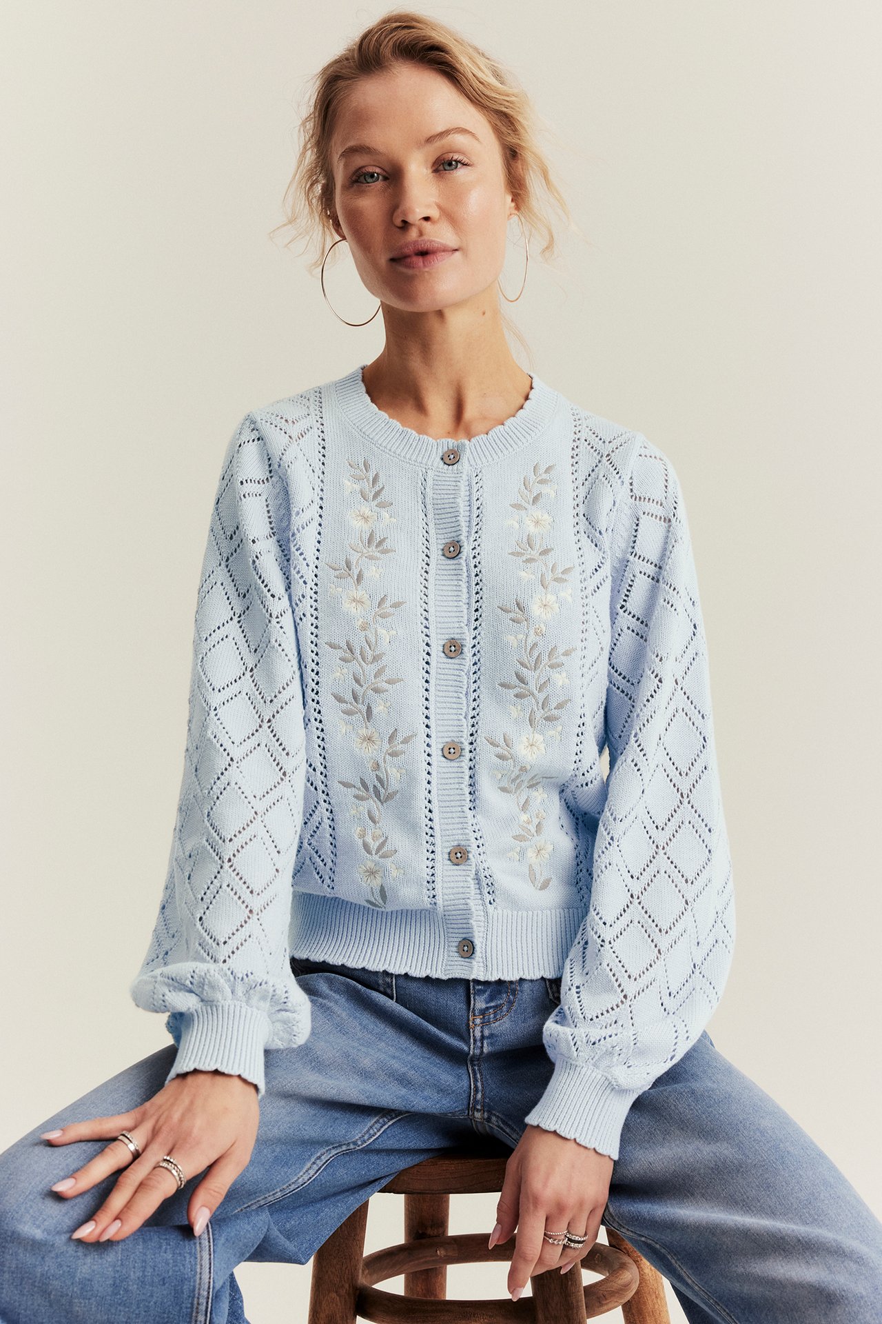 Front view: Woman wears a light blue embroidered cotton knitted cardigan with blue jeans.