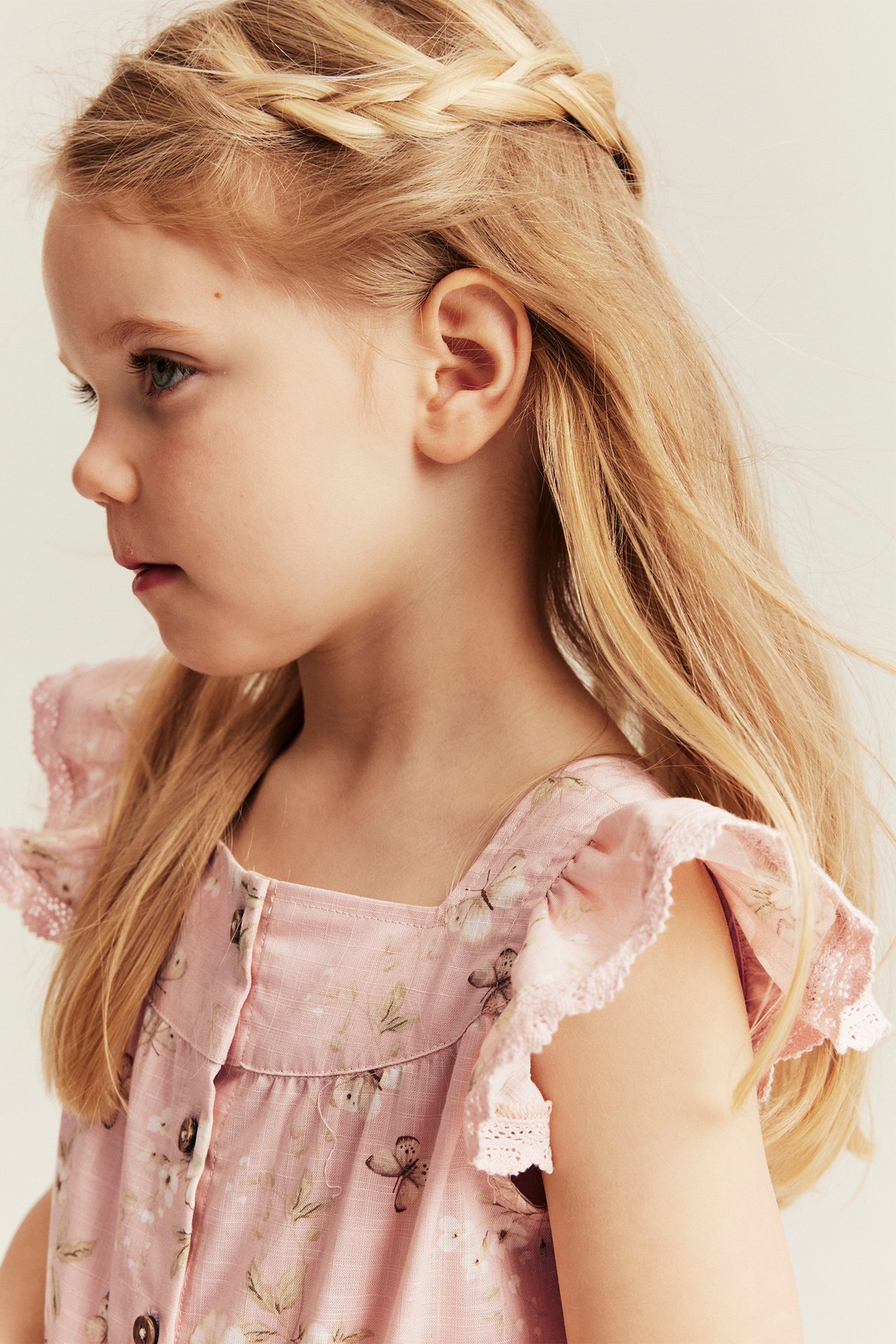 Close-up side view of a kid wearing a light pink floral jumpsuit with ruffled, lace-trimmed sleeves and front buttons.