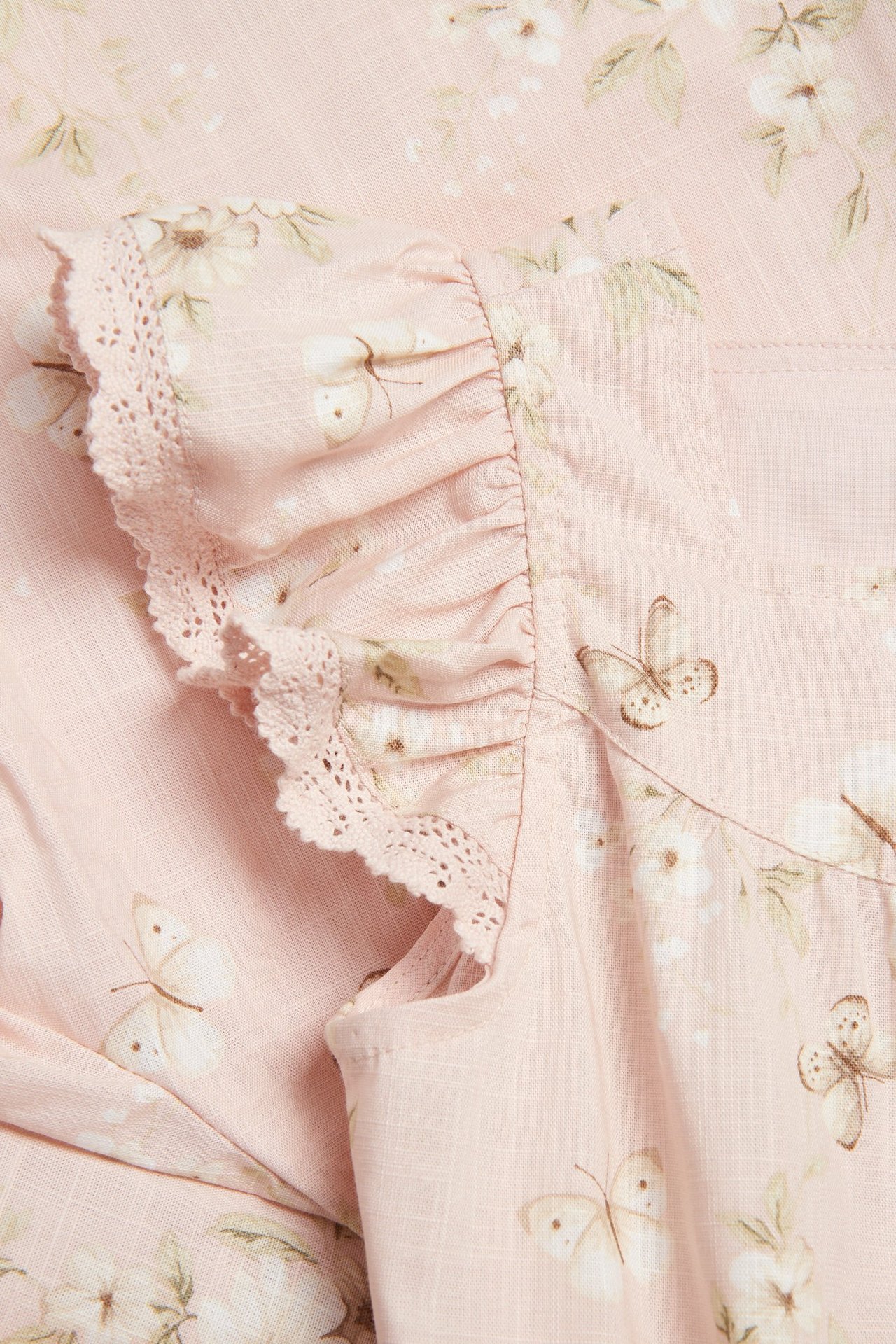 Nahaufnahme eines hellrosa gewebten Overalls mit Blumen- und Schmetterlingsmuster, Rüschenärmel mit Spitzenbesatz.