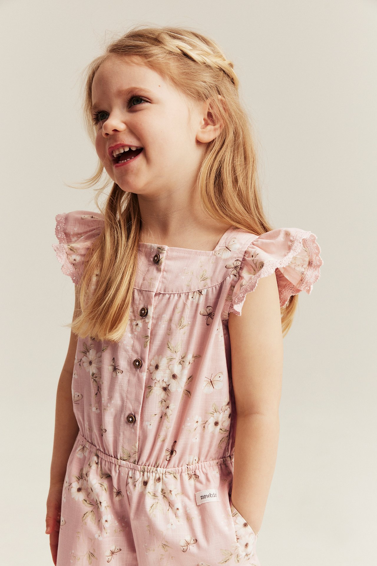 Vorderansicht: Kind in hellrosa Baumwoll-Jumpsuit mit Blumenmuster, Rüschen-Spitzenärmeln.