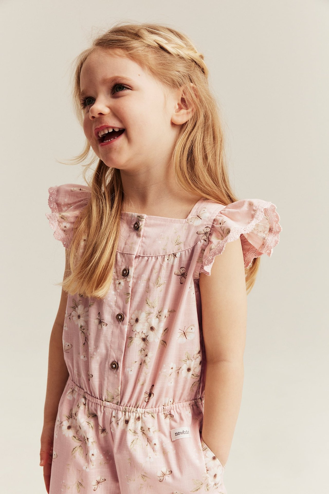 Vorderansicht: Kind in hellrosa Baumwoll-Jumpsuit mit Blumenmuster, Rüschen-Spitzenärmeln.