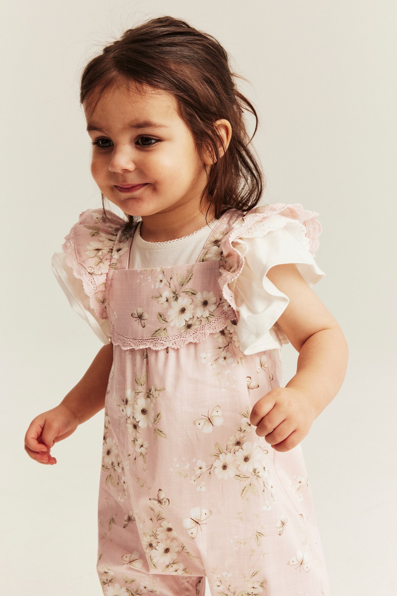 Vorderansicht: Baby-Mädchen in einem hellrosa geblümten Baumwoll-Jumpsuit mit Rüschenärmeln, über einem weißen Oberteil.