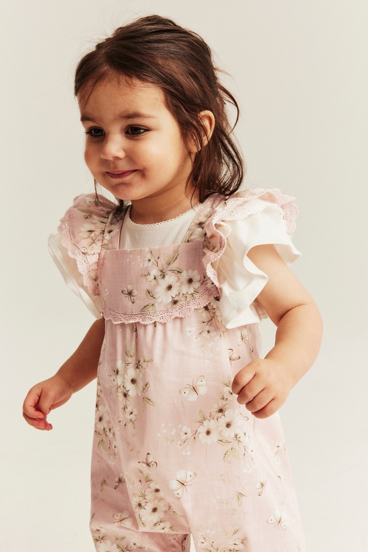 Vorderansicht: Baby-Mädchen in einem hellrosa geblümten Baumwoll-Jumpsuit mit Rüschenärmeln, über einem weißen Oberteil.