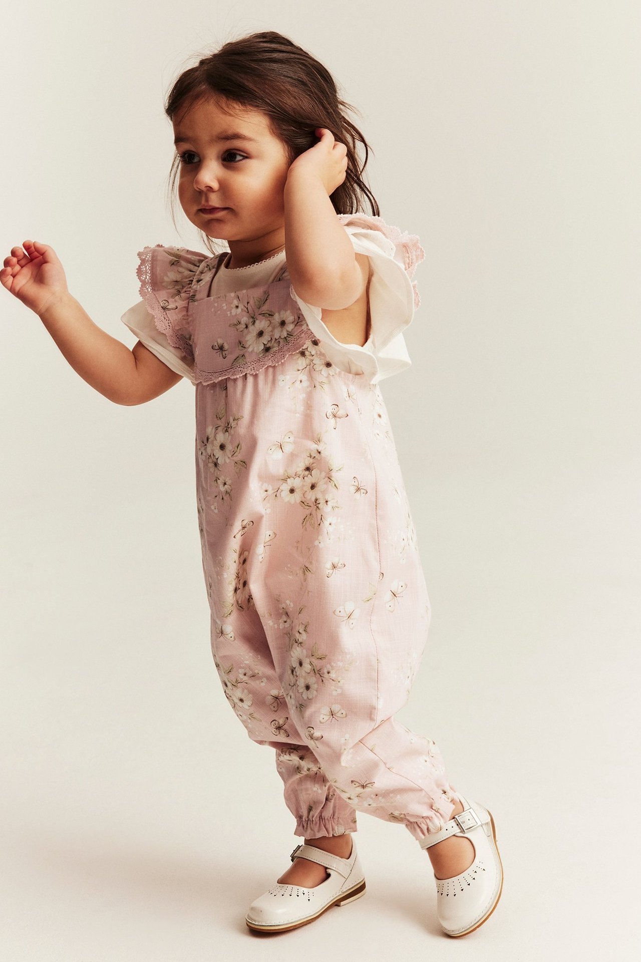Vorderansicht: Kind in hellrosa geblümtem Overall mit Rüschen, über einem weißen T-Shirt und weißen Schuhen.