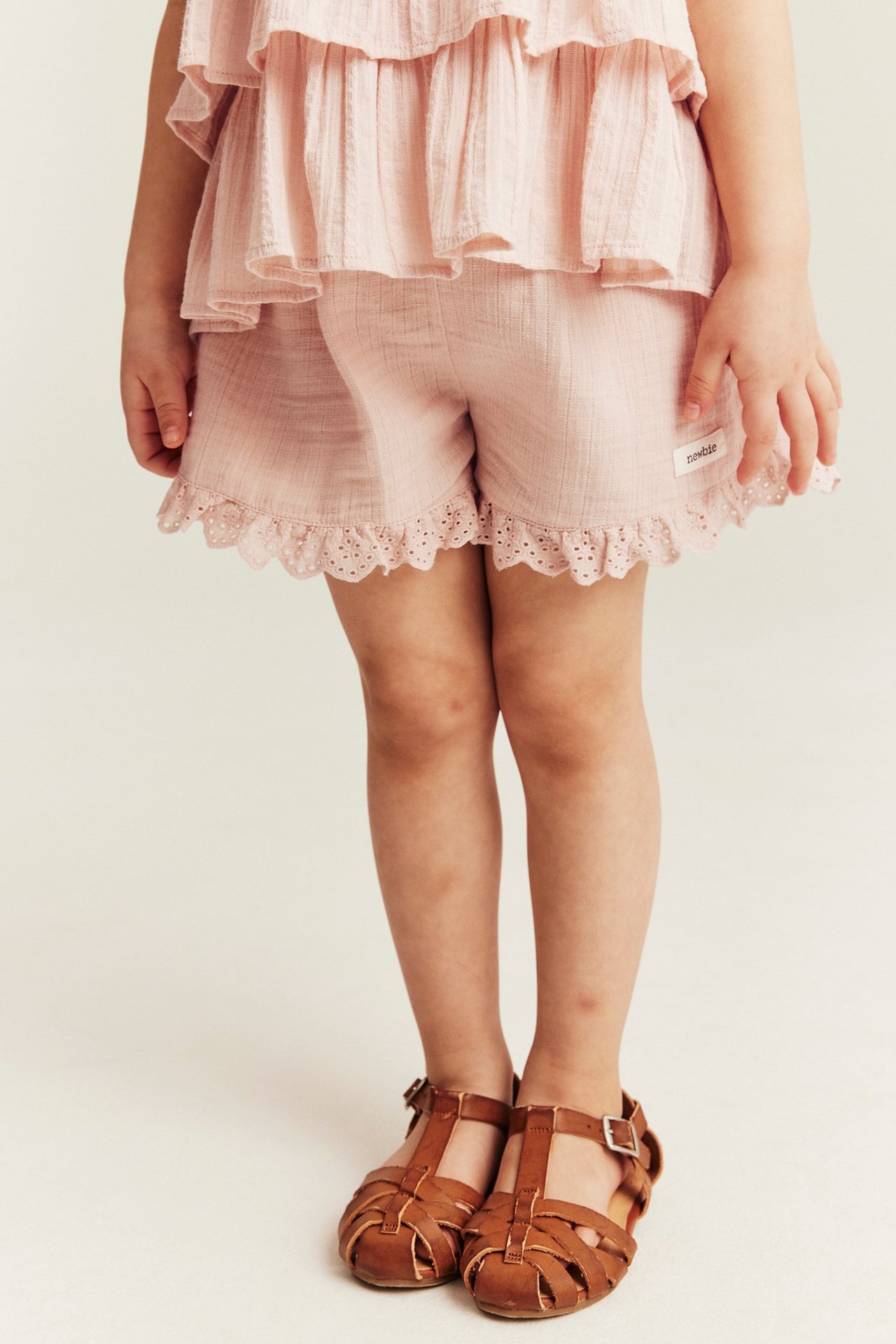 Front view: Kid wears light pink woven shorts with lace trim, a ruffled top, and brown sandals.