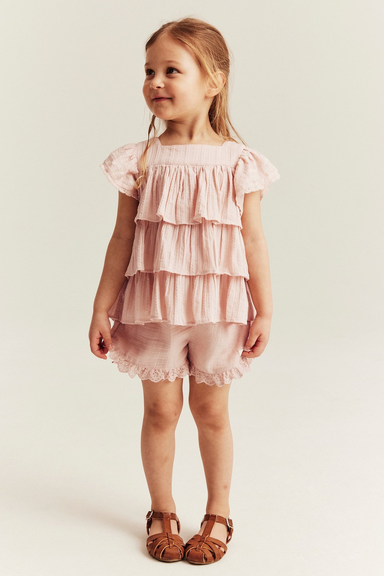 Front view of a kid wearing light pink woven shorts with lace trim, a tiered top, and brown sandals.