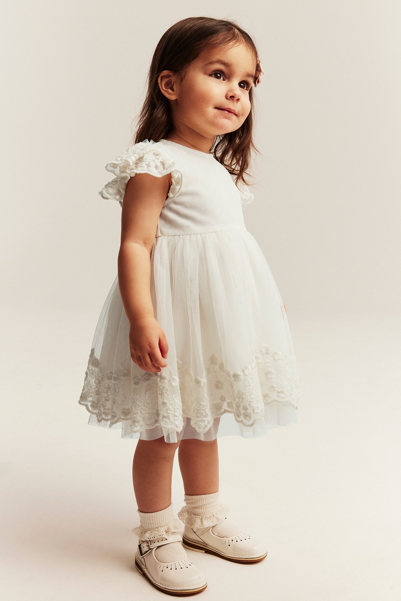 Seitenansicht, kleines Mädchen im cremefarbenen bestickten Netz-Kleid mit Rüschenschultern, Rüschensocken und weißen Mary Jane Schuhen.