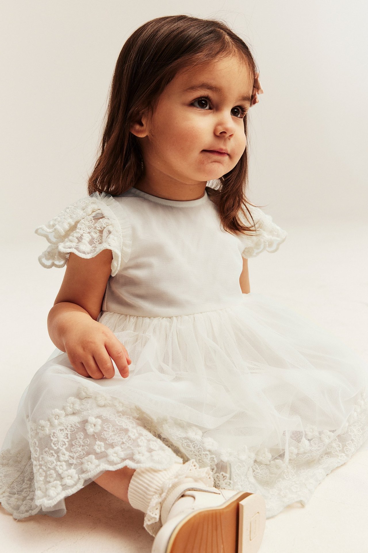 A sitting baby girl wears an off-white embroidered mesh dress with ruffled sleeves, ribbed socks, and white shoes.