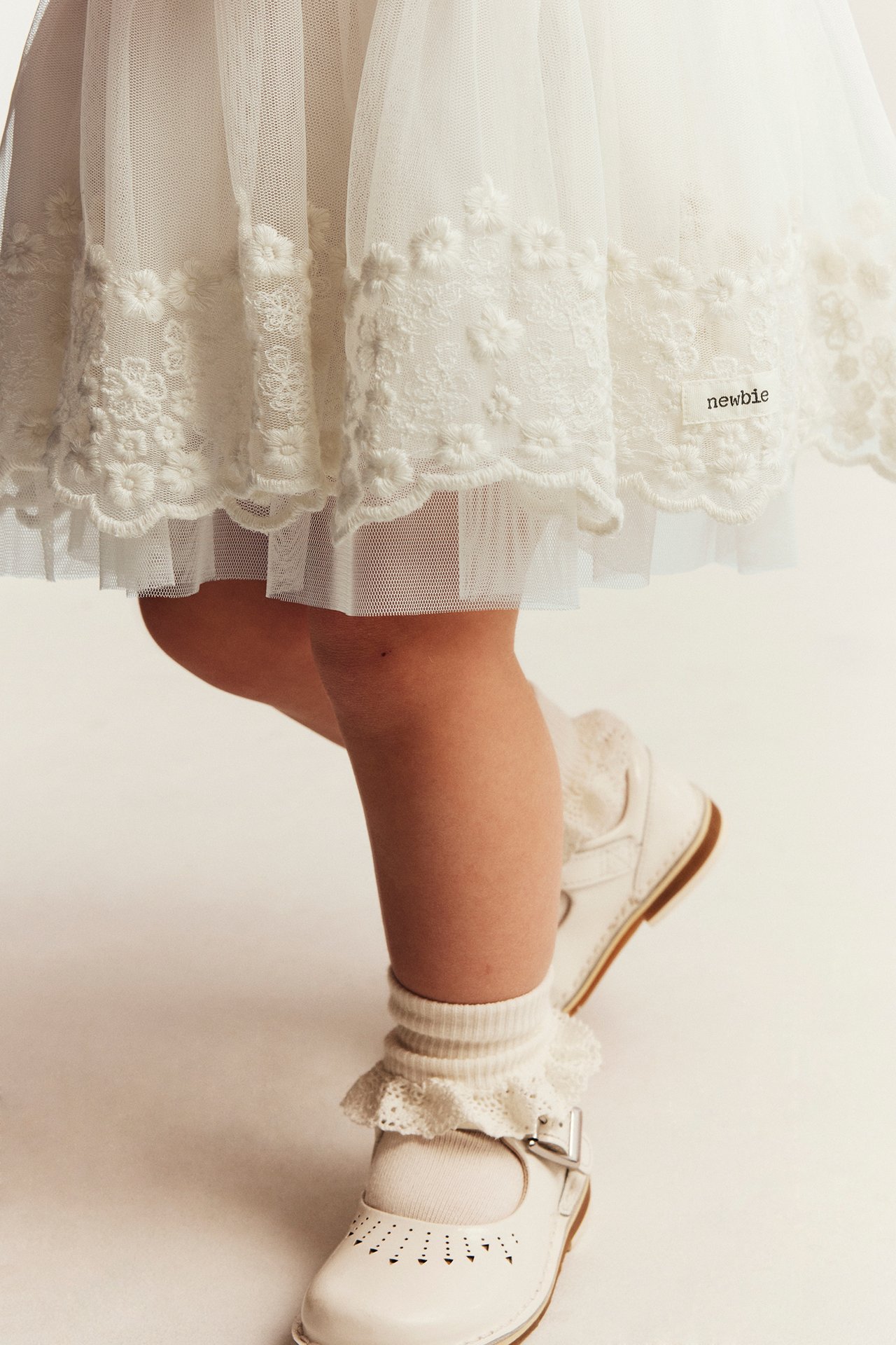 Close-up of a kid's offwhite mesh dress with floral embroidery, worn with white frilled socks and Mary Jane shoes.