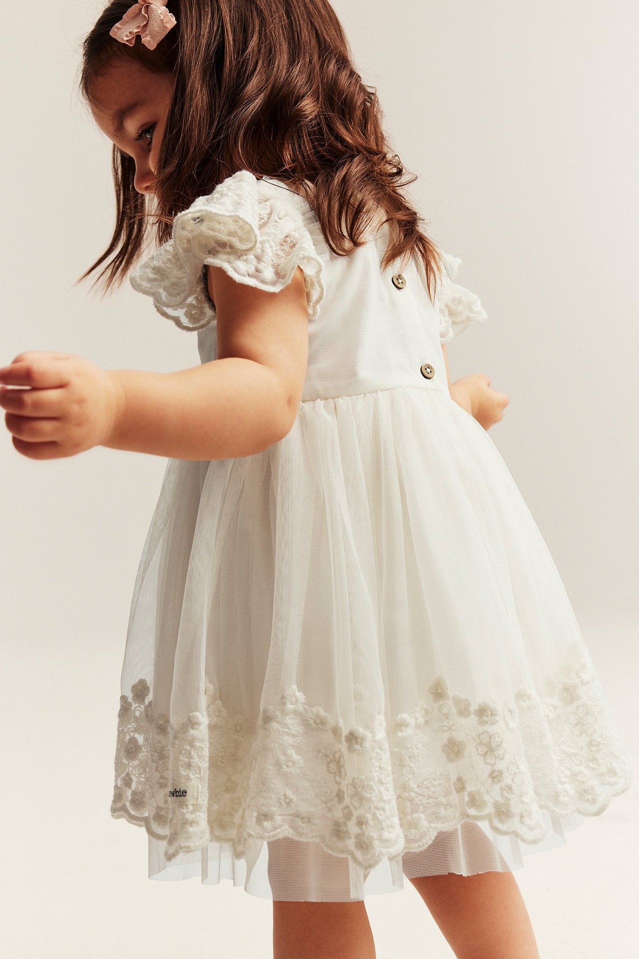 Kind in cremeweißem, besticktem Mesh-Kleid, Rüschenärmeln und Rückenknöpfen, Seiten-Rückenansicht.