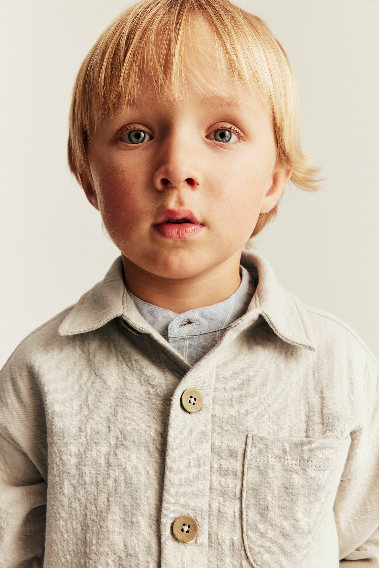 Nahaufnahme, Vorderansicht eines Kindes in beigem Overshirt mit Knöpfen über hellblauem Ringelhemd.