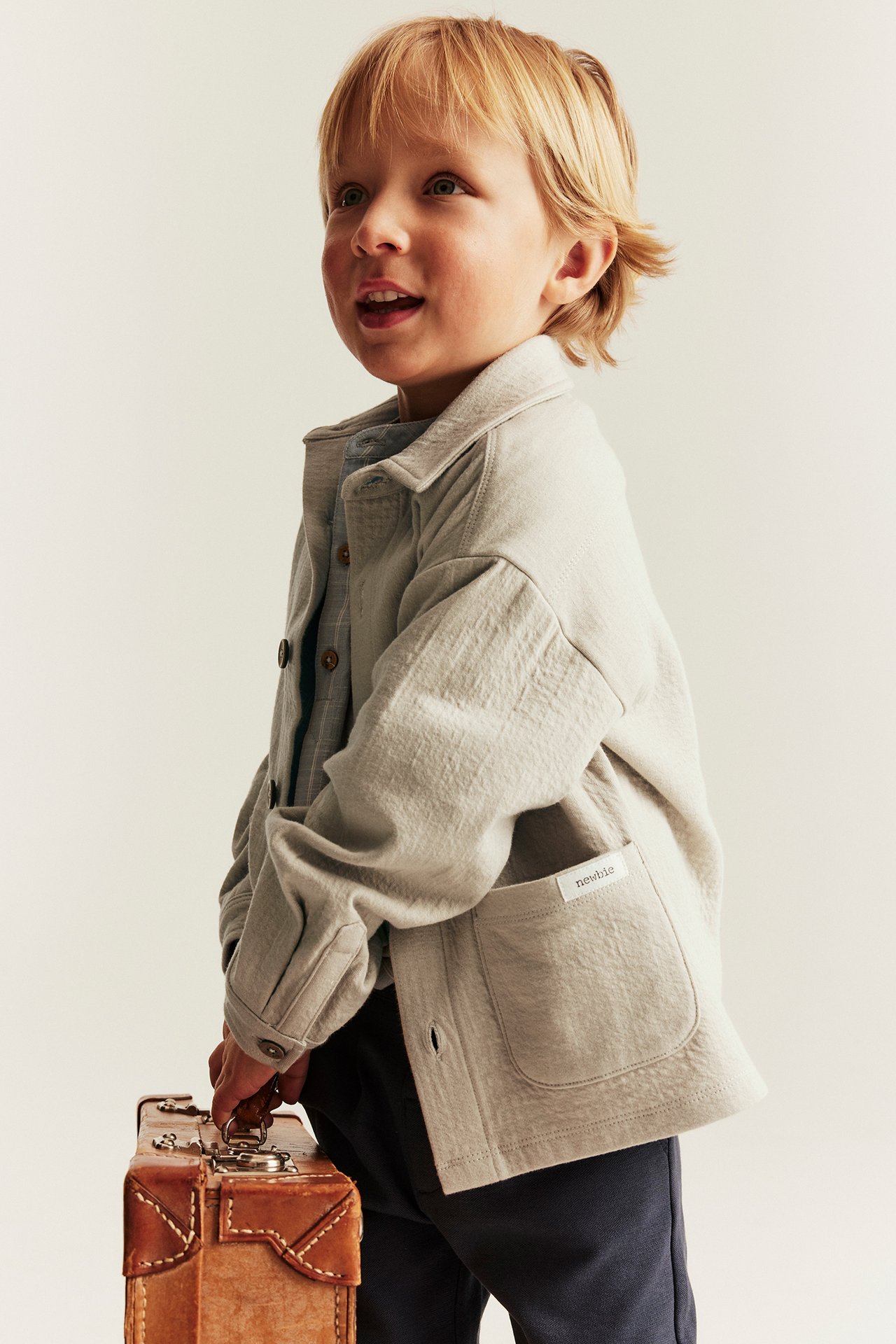 Kind in beigem Kragen-Overshirt über blauem Hemd, dunkler Hose, einen braunen Koffer haltend.