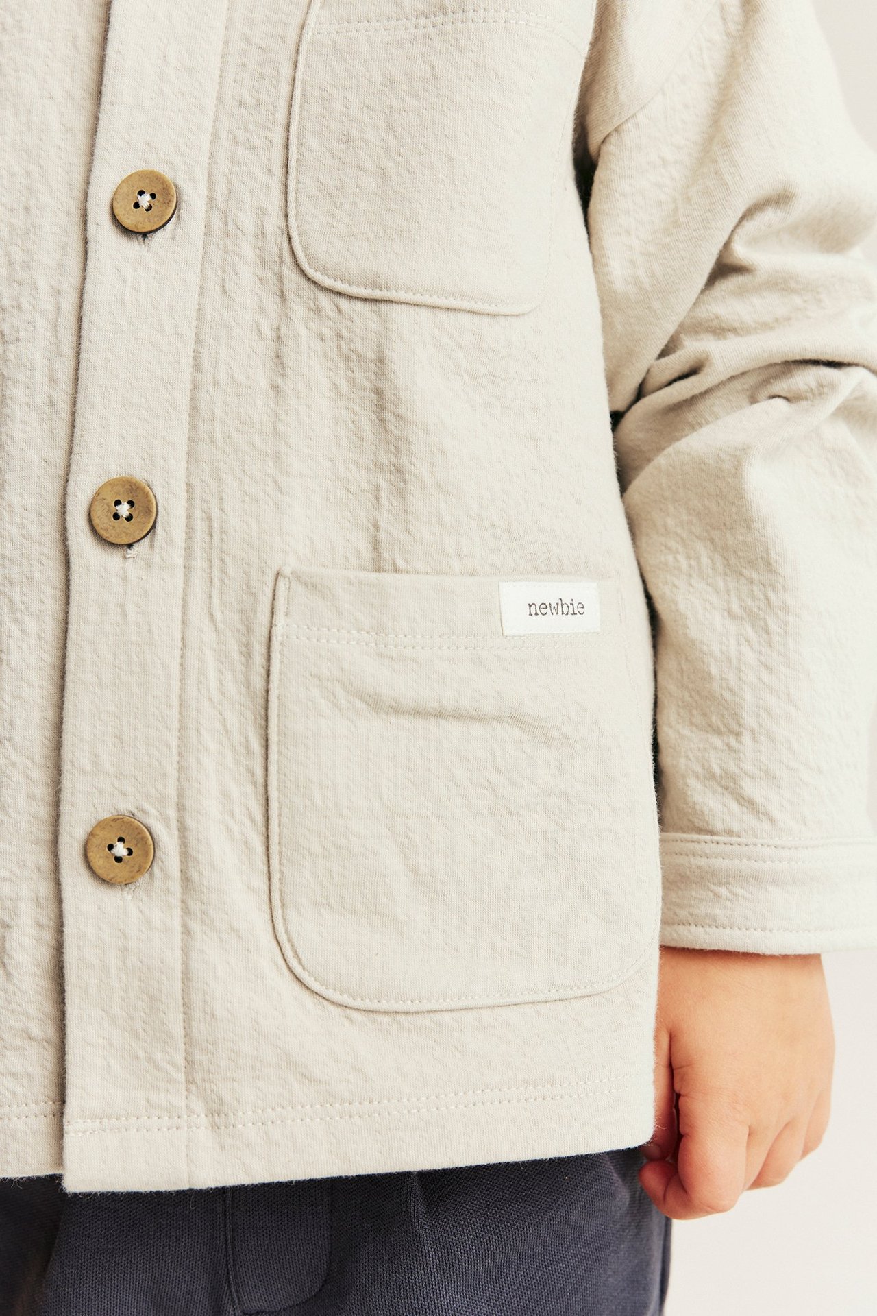 Nahaufnahme eines Kindes in beiger strukturierter Strick-Overshirt mit Kragen, Holzknöpfen und Vordertaschen.