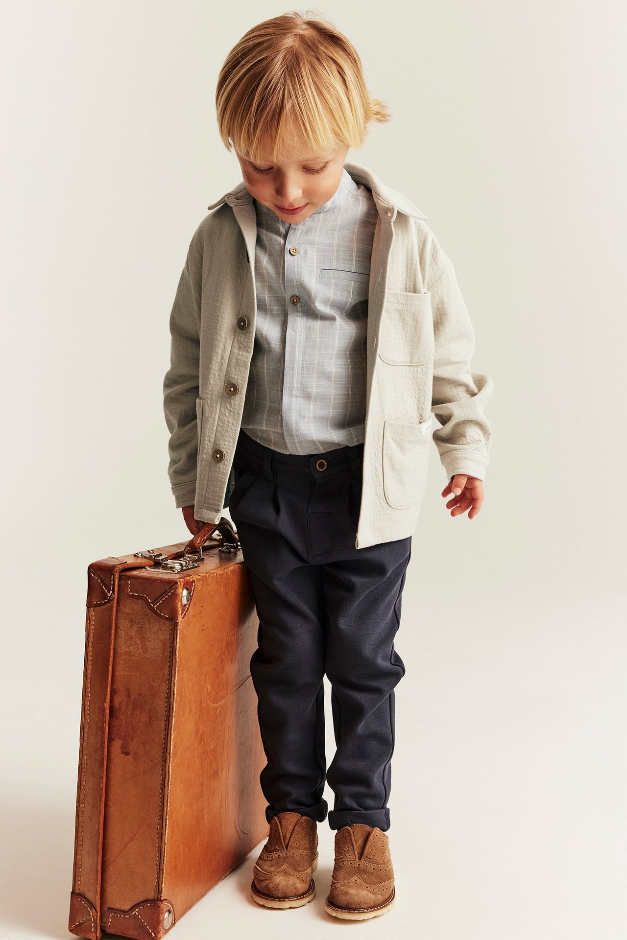 Kid wears beige knit overshirt, plaid shirt, dark blue trousers, brown boots, by a brown suitcase.
