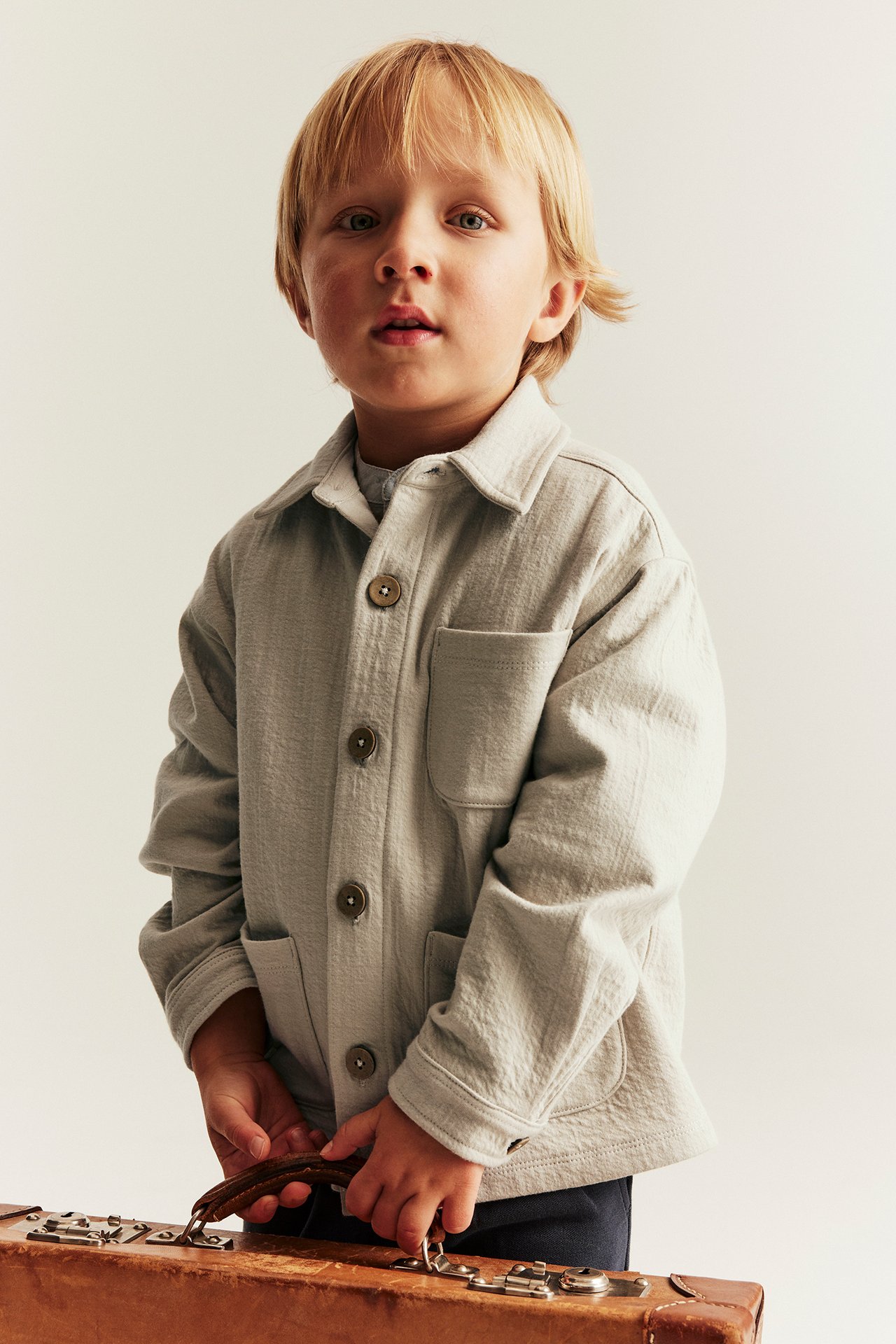 Front view: Kid in a beige structured jersey overshirt with collar, pockets, holding a brown suitcase.