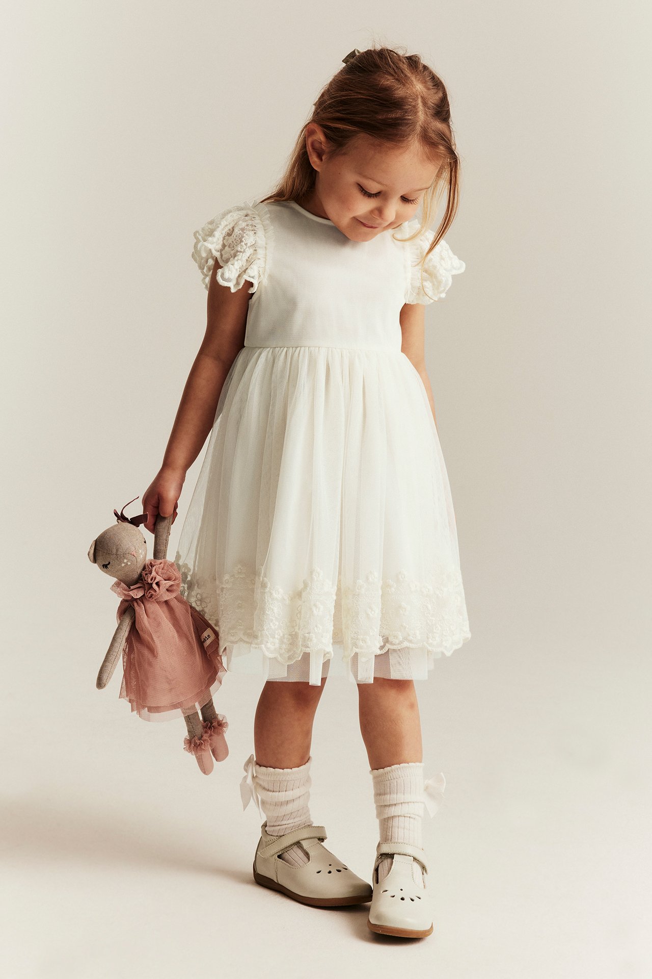 Vorderansicht: Kind in cremefarbenem, besticktem Netz-Kleid, hält Puppe, trägt beige Schuhe und Socken.