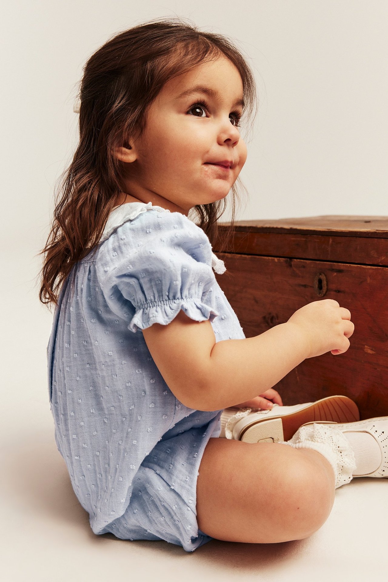 Seitenansicht Baby in hellblauem gepunkteten Playsuit mit weißem Matrosenkragen, weißen Socken und Schuhen.