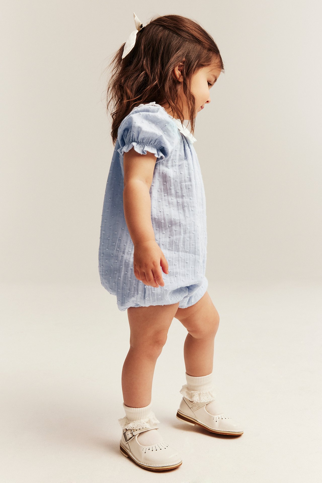 Seitenansicht: Baby in hellblauem Tupfen-Playsuit mit weißem Matrosenkragen, weißen Socken und beigen Spangenschuhen.