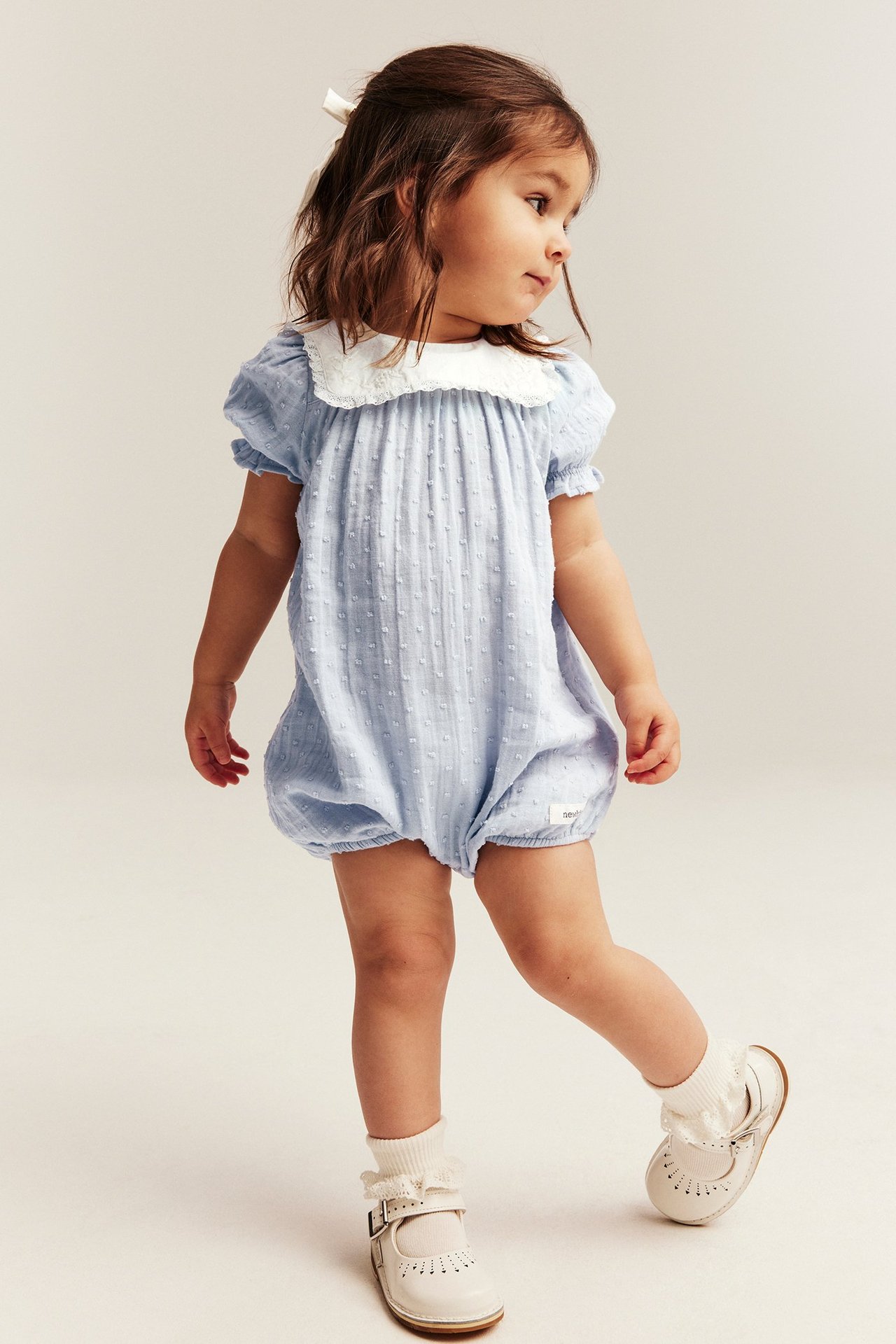 Vorderansicht: Baby in hellblauem Tupfen-Playsuit mit weißem Spitzenkragen und weißen Mary Jane Schuhen.