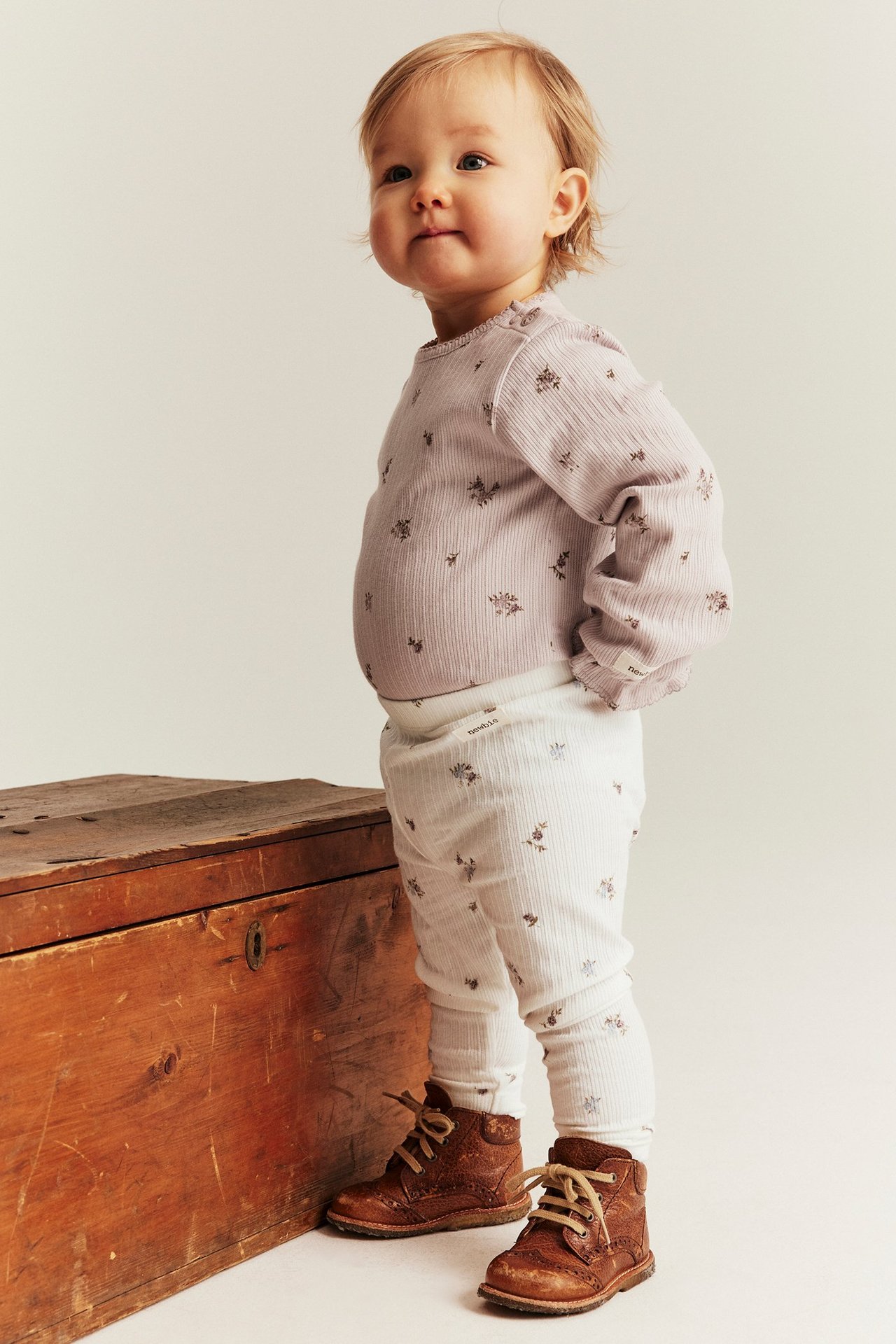 Seitenansicht eines Babys in offwhite gerippten floralen Leggings, einem lila gemusterten Top und braunen Stiefeln.