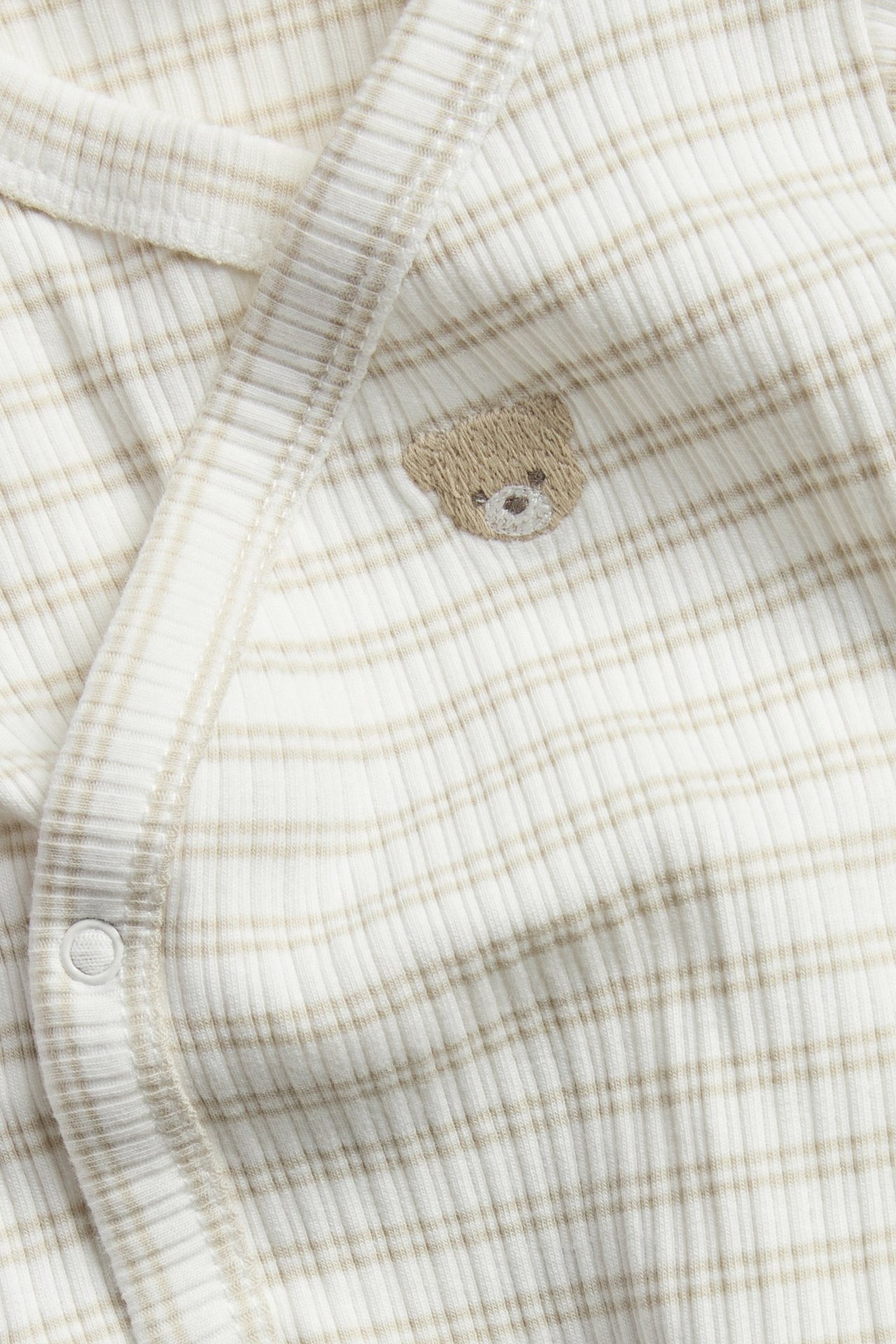 Nahaufnahme eines beigen, gerippten Baby-Wickelbodys mit Streifen und einem kleinen bestickten Bären.