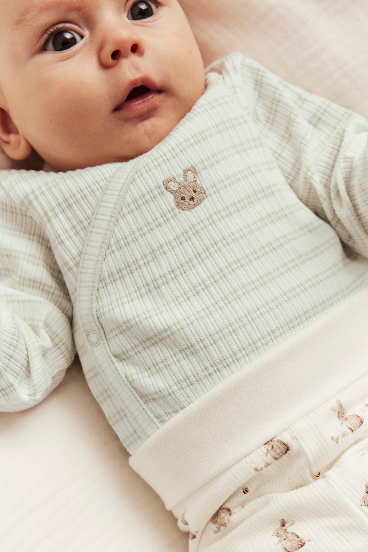 Close-up of baby wearing a light green striped ribbed wrap-over body with bunny embroidery and white patterned pants.