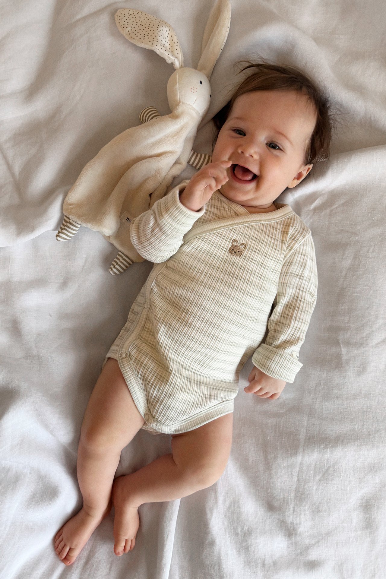 Smiling baby in a light green striped wrap-around bodysuit with an embroidered animal, next to a beige bunny comfort blanket.