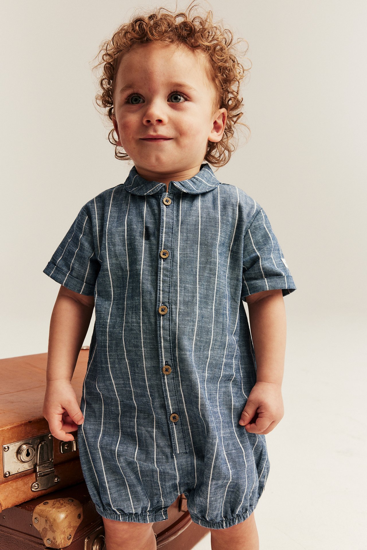 Front view of a baby in a blue striped woven cotton romper with collar and front buttons.
