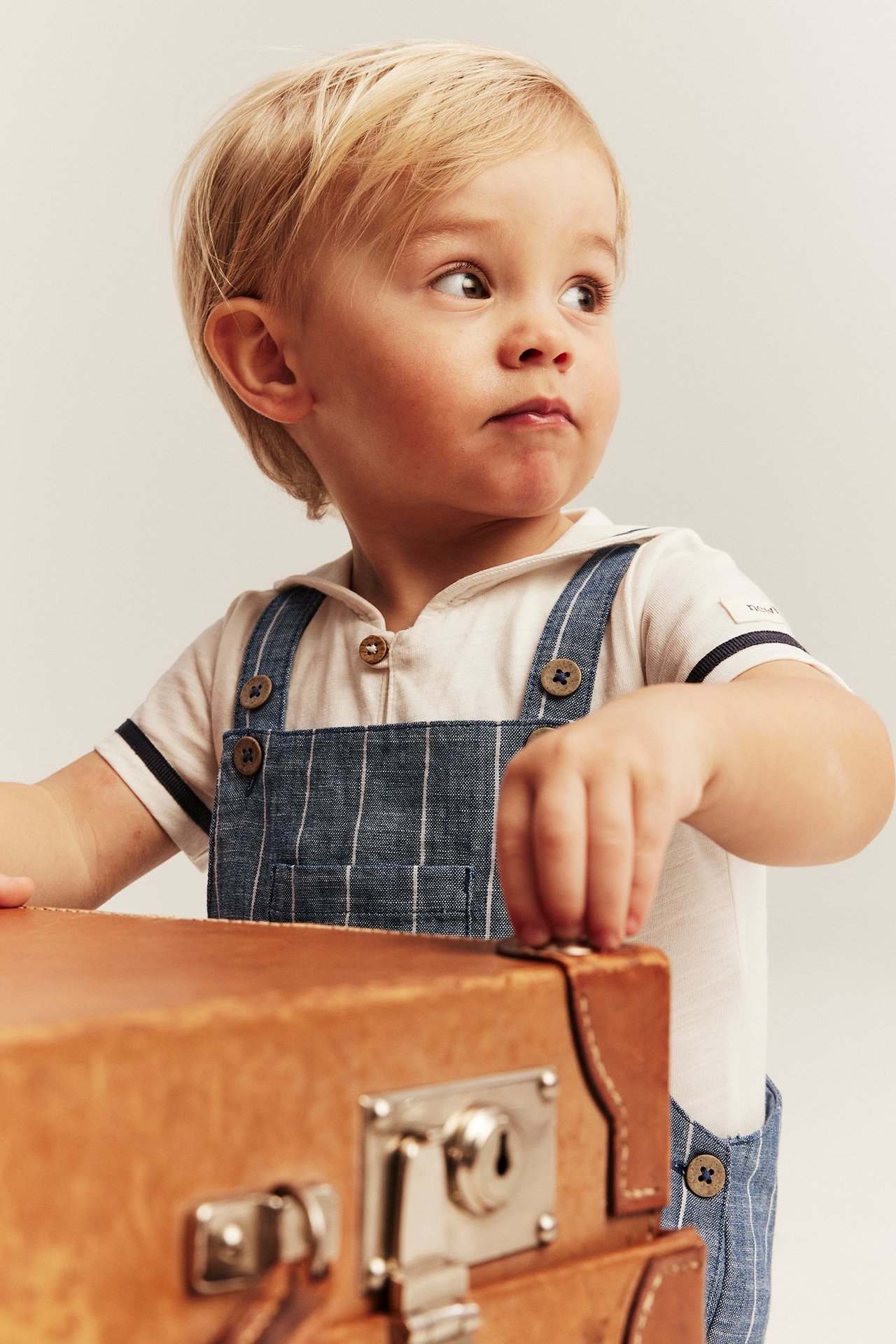 Baby boy wears blue striped overalls with buttons over a cream t-shirt, next to a brown suitcase.