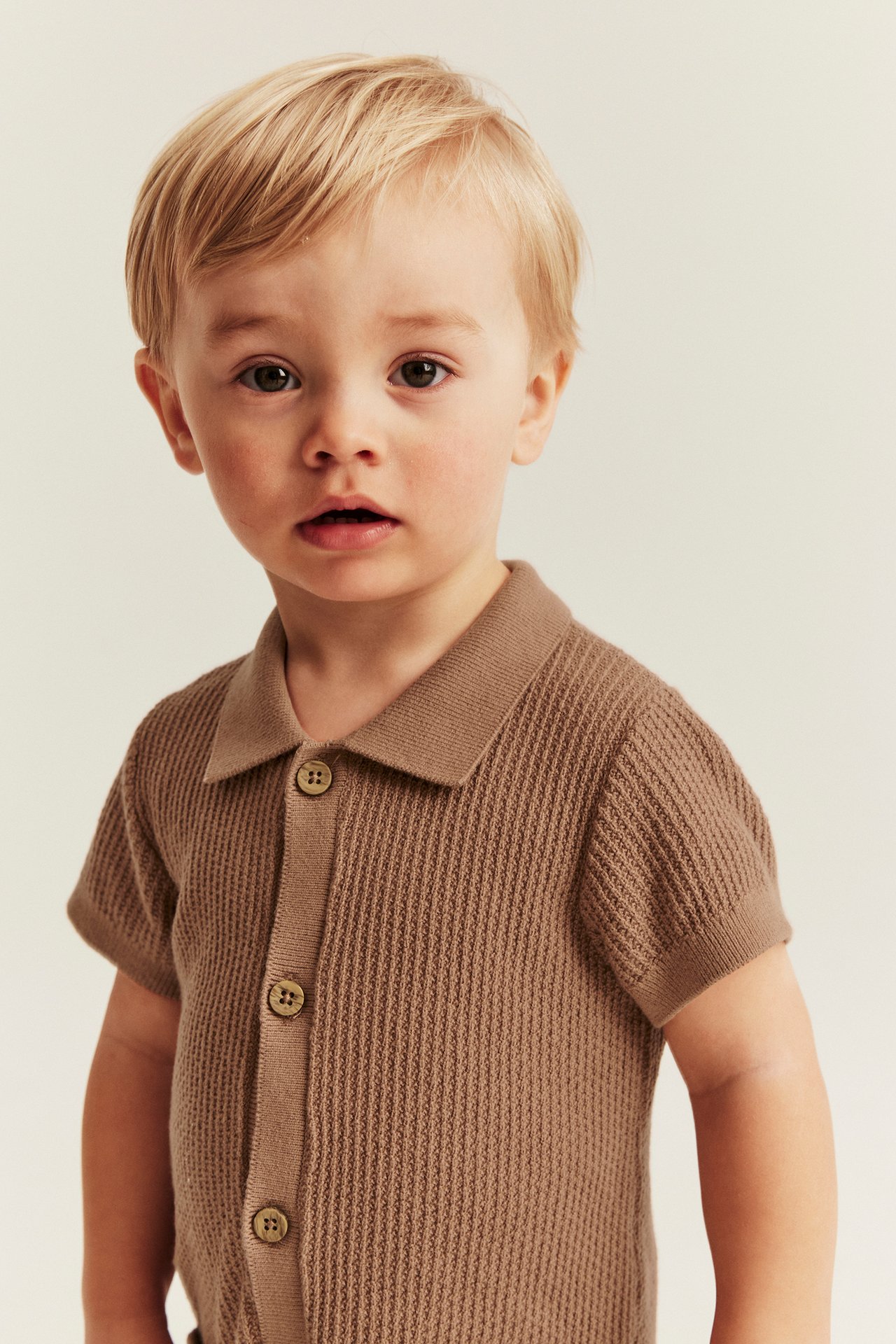 Front view of a baby wearing a brown finely knitted short-sleeved romper with a collar and front buttons.