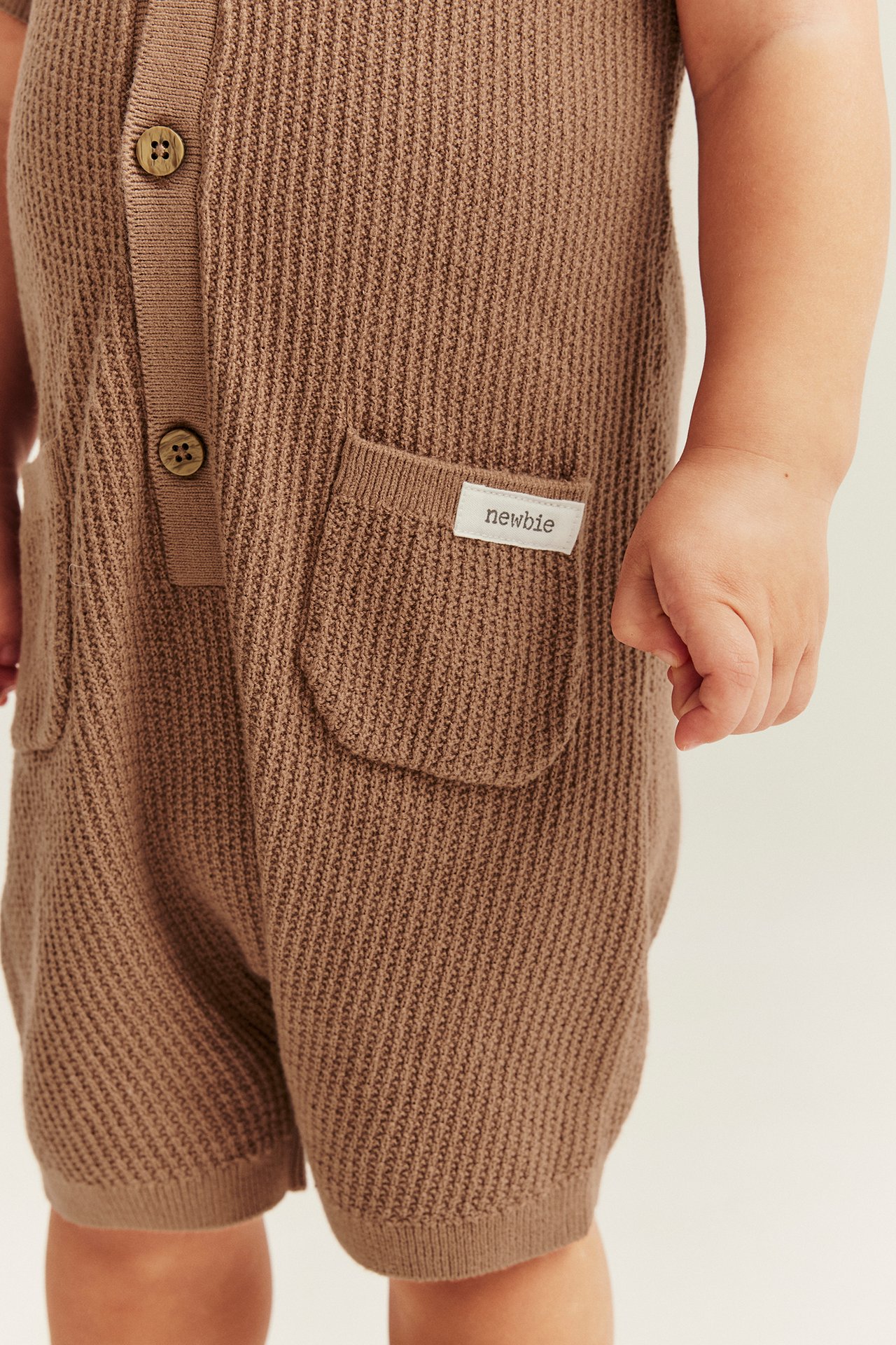 Close-up front view of a baby wearing a brown finely knitted cotton romper with front buttons and pockets.