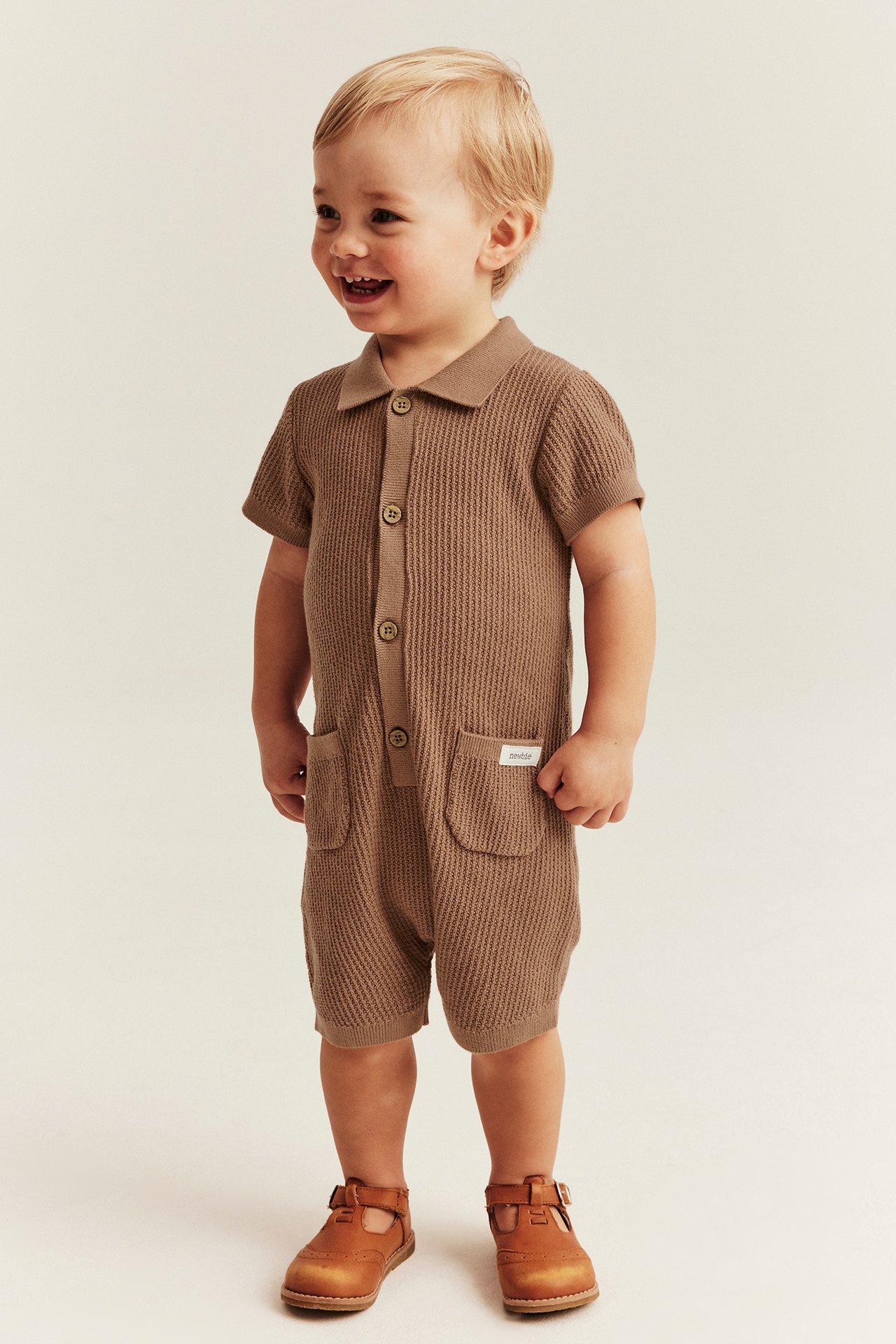 Front view of a laughing baby wearing a brown finely knitted short-sleeved romper with two pockets and brown T-bar shoes.
