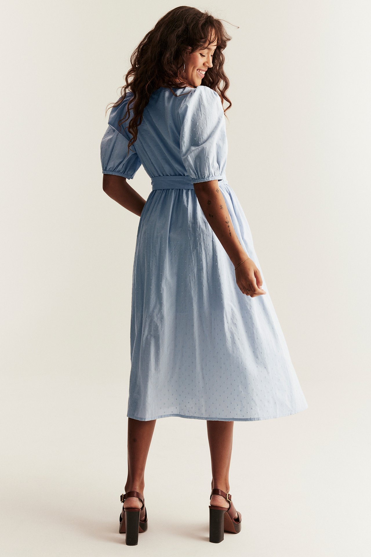 Woman, back view: light blue cotton midi dress with puff sleeves, tie-belt, and brown block-heel sandals.