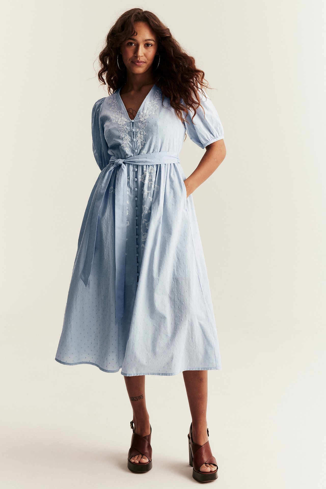 Woman in light blue cotton midi dress with white embroidered flowers, tie belt, and puff sleeves. Front view.