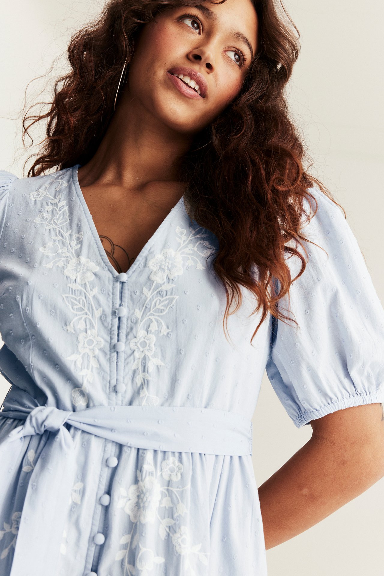 Close-up of a woman in a light blue cotton dress with white floral embroidery, buttons, and tied belt.