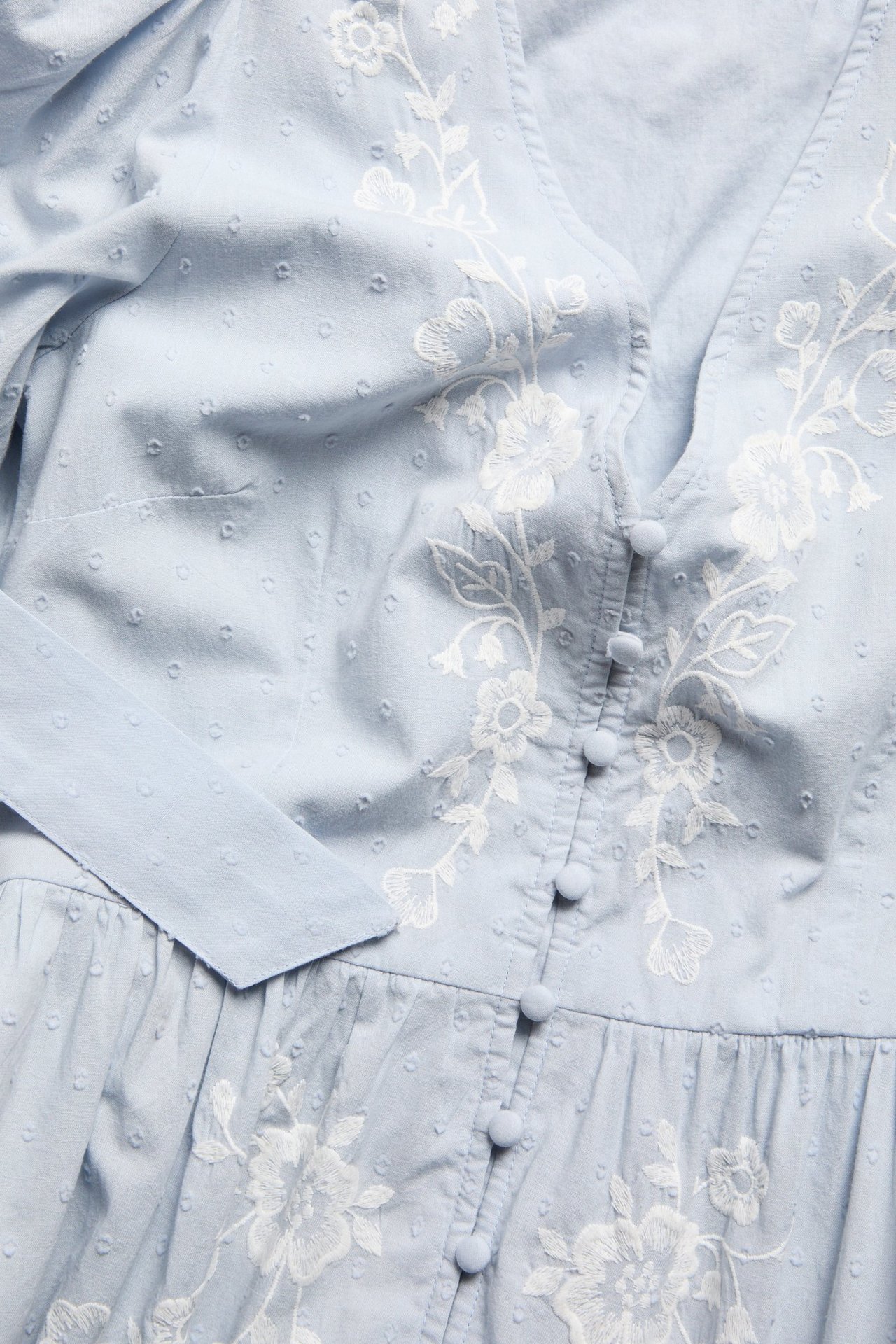 Close-up front view of a light blue cotton dress with white embroidered flowers, textured dots, and covered buttons.